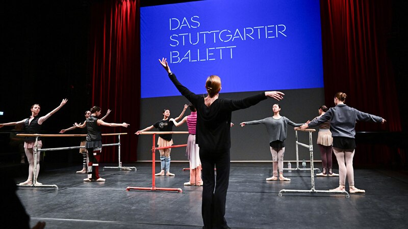 Ballet rehearsal with dancers and trainer in front of a screen with the inscription 'Das Stuttgarter Ballett'., &copy; W&uuml;rttembergische Staatstheater Stuttgart