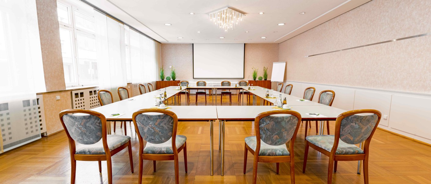 Heller Konferenzraum mit U-förmiger Tischanordnung, Stühlen, Leinwand und Flipchart. Elegante Beleuchtung und Holzboden., © TOMAS