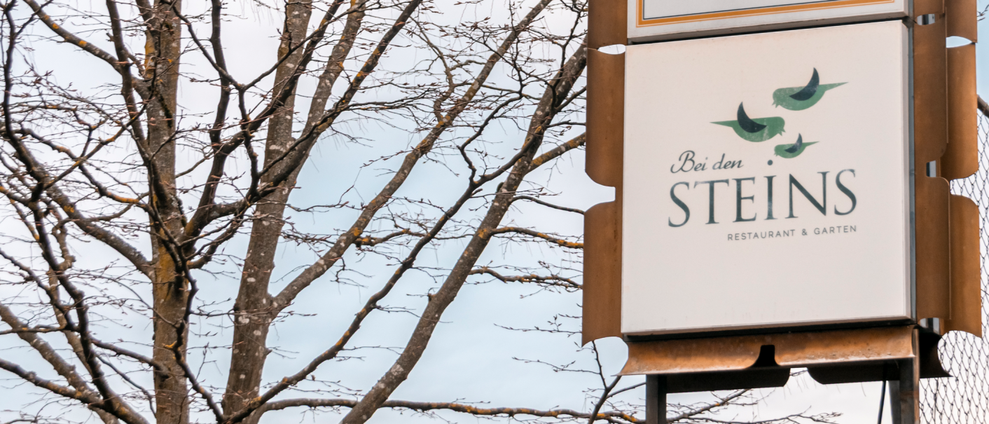 Ein Schild des Restaurants 'Bei den Steins' mit V&ouml;geln als Logo, vor einem kahlen Baum und blauem Himmel., &copy; Stuttgart-Marketing GmbH, Sarah Schmid