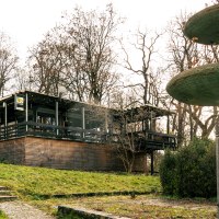 Gebäude mit Holzverkleidung und Terrasse in bewaldeter Umgebung, daneben ein großer, mehrstufiger Brunnen. Sonnenstrahlen scheinen durch die Bäume., © Stuttgart Marketing GmbH, Sarah Schmid Gebäude mit Holzverkleidung und Terrasse in bewaldeter Umgebung, daneben ein großer, mehrstufiger Brunnen. Sonnenstrahlen scheinen durch die Bäume., © Stuttgart Marketing GmbH, Sarah Schmid