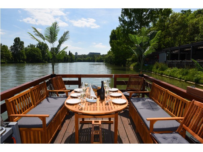Ein Floß mit Holzmöbeln und gedecktem Tisch auf einem ruhigen Fluss, umgeben von Bäumen., © Cool-Tours StattReisen Ein Floß mit Holzmöbeln und gedecktem Tisch auf einem ruhigen Fluss, umgeben von Bäumen., © Cool-Tours StattReisen