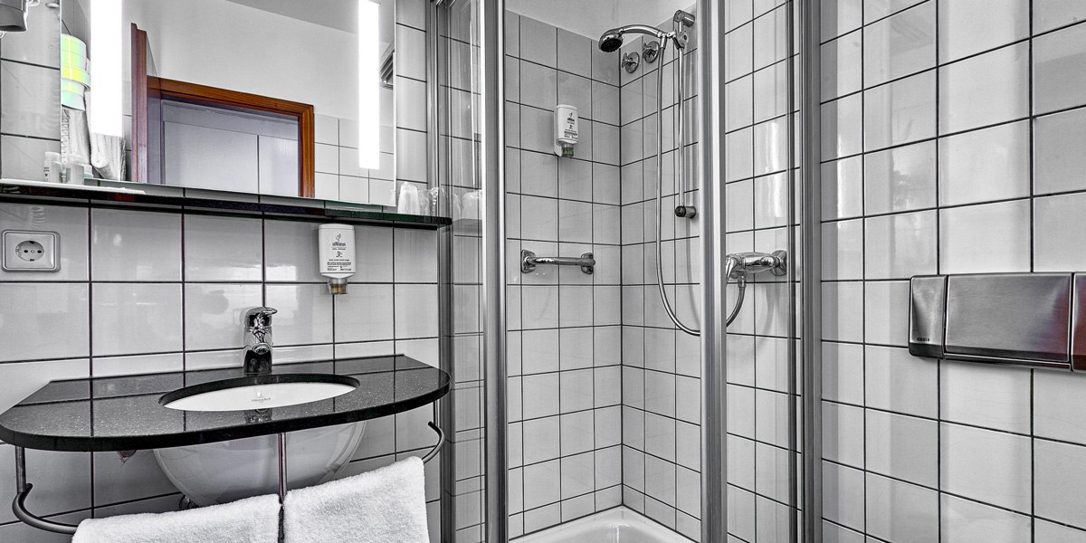 Modernes Badezimmer mit weiß gefliesten Wänden, Duschkabine, Waschbecken mit schwarzer Ablage und beleuchtetem Spiegel., © attimo Modernes Badezimmer mit weiß gefliesten Wänden, Duschkabine, Waschbecken mit schwarzer Ablage und beleuchtetem Spiegel., © attimo