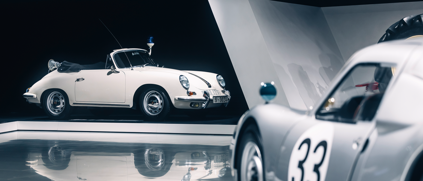 Two classic cars in an exhibition. A white convertible with police lights and a silver car with the number 33 in the foreground., © Porsche AG Two classic cars in an exhibition. A white convertible with police lights and a silver car with the number 33 in the foreground., © Porsche AG