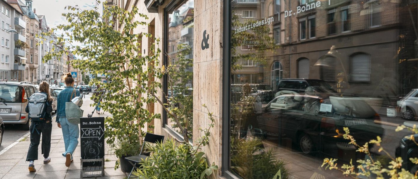 Zwei Personen gehen an einem Geschäft mit der Aufschrift 'Dr. Bohne' vorbei. Die Straße ist von Autos gesäumt, und Pflanzen stehen vor dem Schaufenster., © SMG, Sarah Schmid