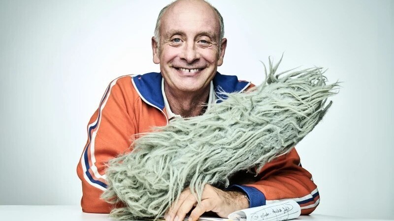 Ein Mann in einer orangefarbenen Jacke lächelt und hält ein flauschiges Mikrofon. Vor ihm liegt ein Notizblock., © Theaterhaus Stuttgart e.V. Ein Mann in einer orangefarbenen Jacke lächelt und hält ein flauschiges Mikrofon. Vor ihm liegt ein Notizblock., © Theaterhaus Stuttgart e.V.