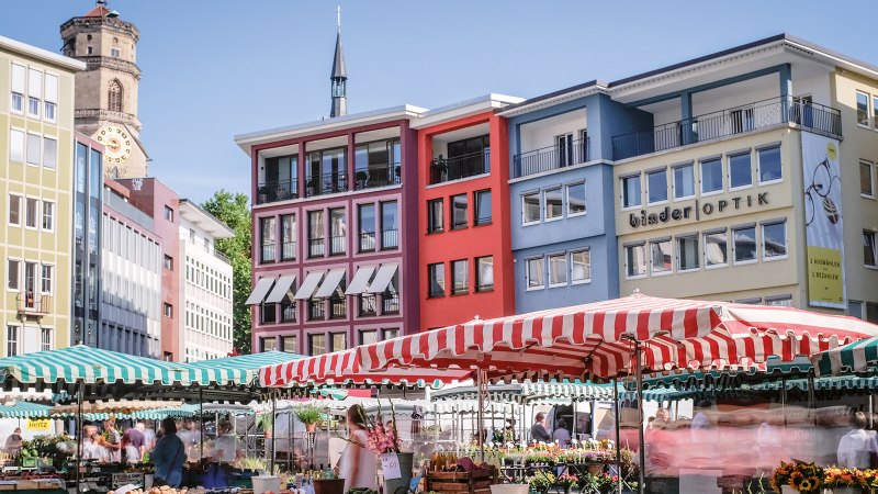 Wochenmarkt in Stuttgart mit bunten Marktständen und Gebäuden im Hintergrund. Menschen schlendern zwischen den Ständen, während die Sonne scheint., © SMG, trickytine