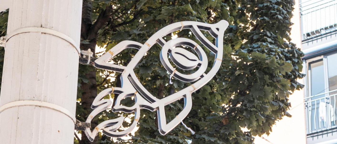 Neon sign in the shape of a rocket with a coffee bean, mounted on a tree in front of a building., © SMG Stuttgart Marketing GmbH - Sarah Schmid Neon sign in the shape of a rocket with a coffee bean, mounted on a tree in front of a building., © SMG Stuttgart Marketing GmbH - Sarah Schmid