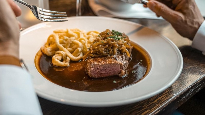 Ein Teller mit Steak, Zwiebeln und Spätzle in Soße. Eine Person hält eine Gabel und ein Messer, bereit zum Essen., © SMG, Martina Denker Ein Teller mit Steak, Zwiebeln und Spätzle in Soße. Eine Person hält eine Gabel und ein Messer, bereit zum Essen., © SMG, Martina Denker