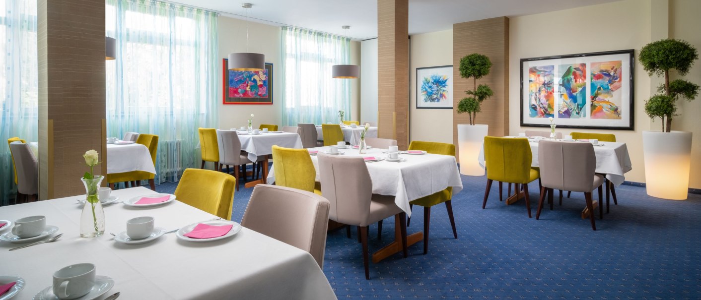 Eleganter Frühstücksraum mit weißen Tischdecken, bunten Stühlen und Kunstwerken an den Wänden. Große Fenster lassen viel Licht herein., © Hetzel Hotel Stuttgart Eleganter Frühstücksraum mit weißen Tischdecken, bunten Stühlen und Kunstwerken an den Wänden. Große Fenster lassen viel Licht herein., © Hetzel Hotel Stuttgart