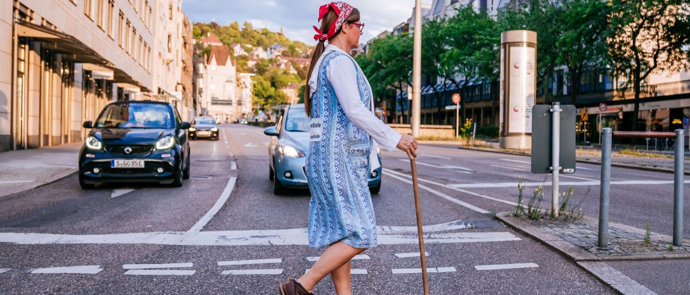 Eine Frau mit Kopftuch und Besen überquert eine Straße in einer Stadt. Autos sind im Hintergrund zu sehen., © Thomas Niedermüller
