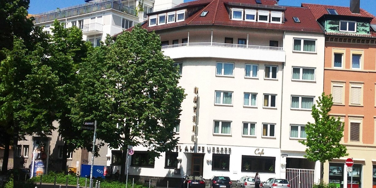 Das Hotel am Feuersee ist ein mehrst&ouml;ckiges Geb&auml;ude mit roten Dachziegeln, umgeben von B&auml;umen und geparkten Autos. Ein Caf&eacute; befindet sich im Erdgeschoss., &copy; ARNOTEL GmbH