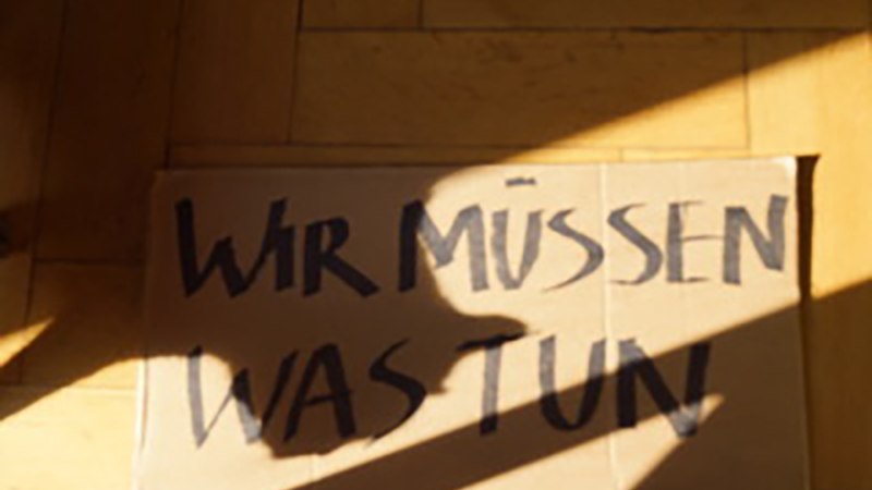 Ein Pappschild mit der Aufschrift 'Wir müssen was tun' liegt auf einem Holzboden, teilweise im Schatten., © Lina Neubeck Ein Pappschild mit der Aufschrift 'Wir müssen was tun' liegt auf einem Holzboden, teilweise im Schatten., © Lina Neubeck