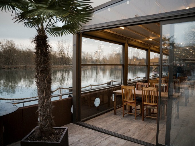 Ein verglaster Wintergarten mit Holztischen und Stühlen bietet einen Blick auf einen ruhigen See. Eine Palme steht im Vordergrund und verleiht dem Ort ein tropisches Flair., © Cool-Tours StattReisen Ein verglaster Wintergarten mit Holztischen und Stühlen bietet einen Blick auf einen ruhigen See. Eine Palme steht im Vordergrund und verleiht dem Ort ein tropisches Flair., © Cool-Tours StattReisen