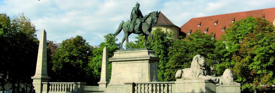 Reiterstatue auf dem Karlsplatz in Stuttgart, flankiert von Löwenstatuen und Obelisken, umgeben von Bäumen und einem historischen Gebäude im Hintergrund., © Stuttgart-Marketing GmbH