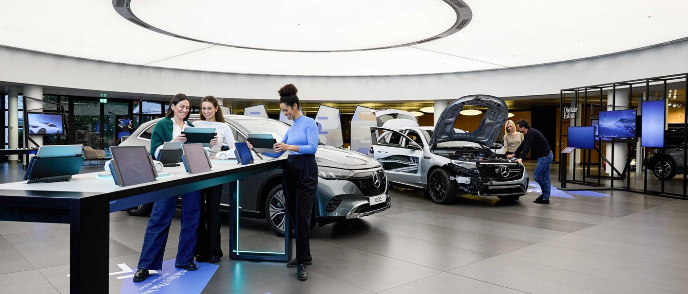 Moderner Ausstellungsraum mit Autos und interaktiven Stationen. Menschen nutzen Tablets, während andere ein Auto mit geöffneter Motorhaube betrachten., © Mercedes-Benz AG Moderner Ausstellungsraum mit Autos und interaktiven Stationen. Menschen nutzen Tablets, während andere ein Auto mit geöffneter Motorhaube betrachten., © Mercedes-Benz AG