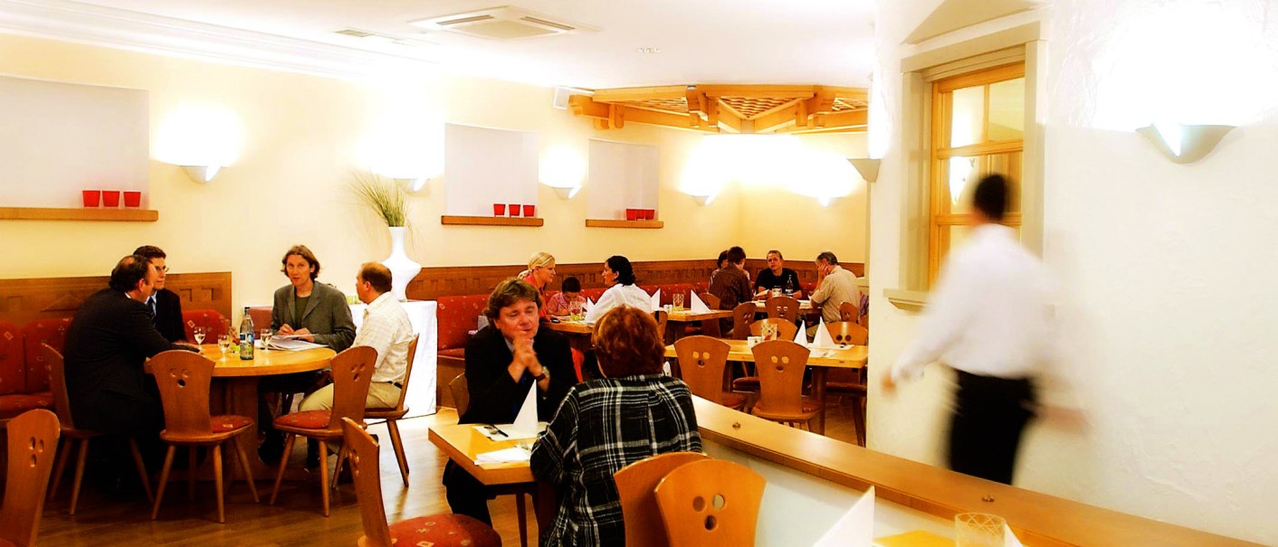Cozy restaurant with wooden furniture, several guests at tables and a waiter on the move. Warm lighting and decorative elements on the walls., © SMG