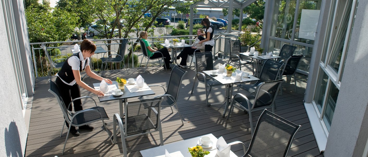 Eine Frühstücksterrasse mit gedeckten Tischen, Blumen und Menschen, die sich unterhalten. Eine Person richtet einen Tisch her., © Hotel Fortuna