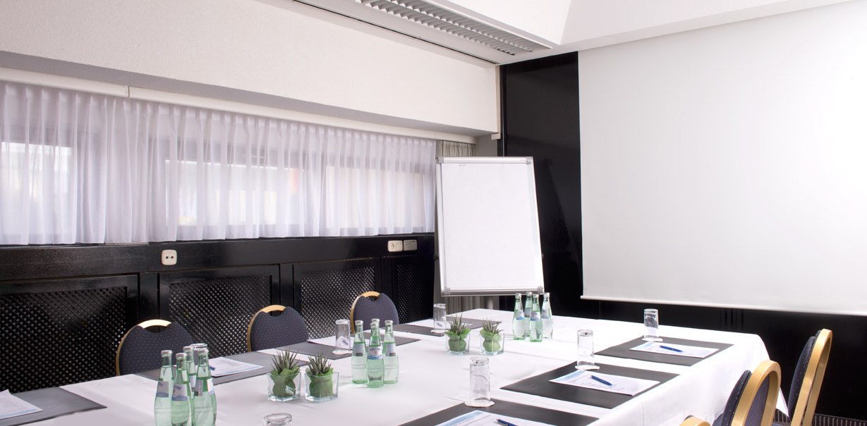 A bright conference room with a table surrounded by chairs. There are water bottles, glasses and writing pads on the table. A flipchart is visible., © Wyndham Stuttgart Airport Messe A bright conference room with a table surrounded by chairs. There are water bottles, glasses and writing pads on the table. A flipchart is visible., © Wyndham Stuttgart Airport Messe