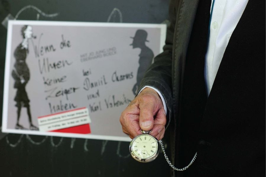 Ein Mann in Anzug hält eine Taschenuhr vor einem Plakat mit dem Titel 'Wenn die Uhren keine Zeiger haben'., © Jim Zimmermann Ein Mann in Anzug hält eine Taschenuhr vor einem Plakat mit dem Titel 'Wenn die Uhren keine Zeiger haben'., © Jim Zimmermann