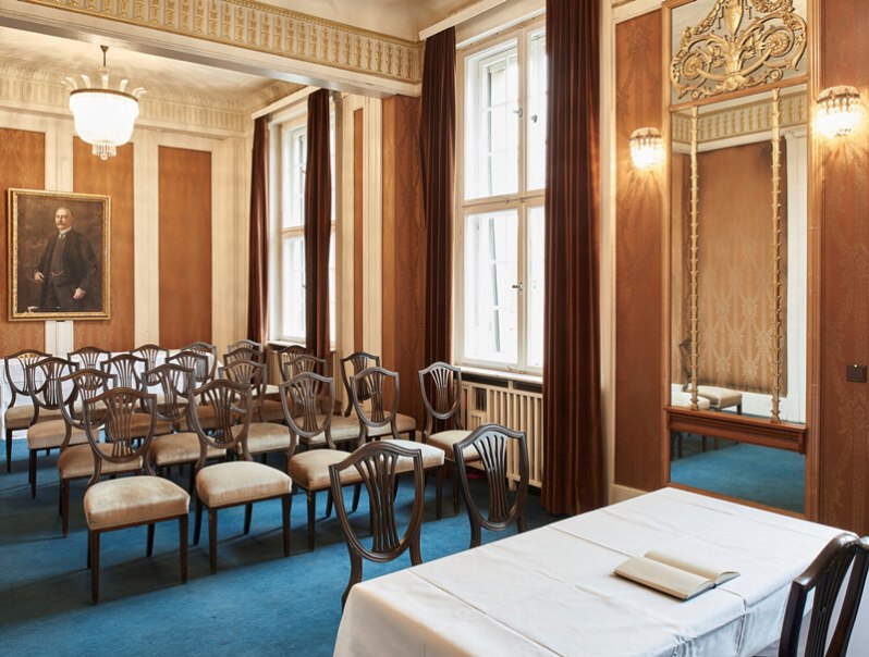 Elegant room with blue carpeting, rows of chairs, a table with a white cloth and a painting on the wall., © Württembergische Staatstheater Stuttgart Elegant room with blue carpeting, rows of chairs, a table with a white cloth and a painting on the wall., © Württembergische Staatstheater Stuttgart
