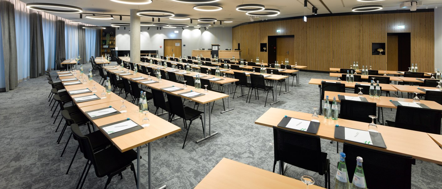Modern conference room with tables, chairs and notepads, illuminated by round ceiling lights. Water bottles and glasses are scattered on the tables., © Holiday Inn Stuttgart Modern conference room with tables, chairs and notepads, illuminated by round ceiling lights. Water bottles and glasses are scattered on the tables., © Holiday Inn Stuttgart