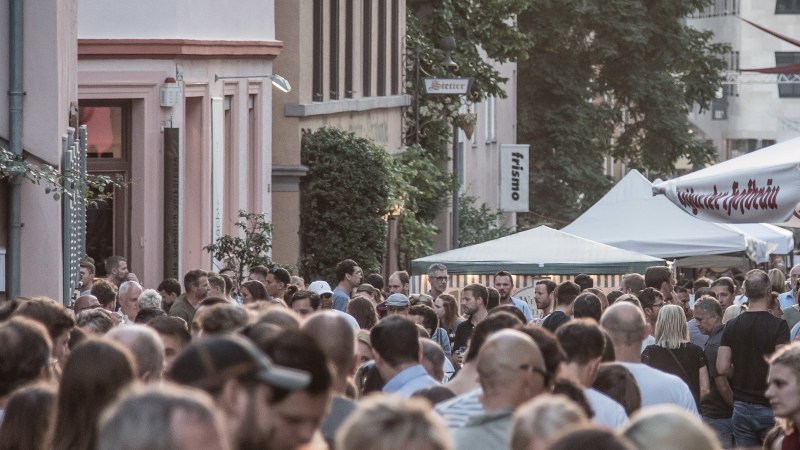 Gro&szlig;e Menschenmenge auf einem Stra&szlig;enfest, umgeben von Geb&auml;uden und Zelten. Im Hintergrund sind Schilder und B&auml;ume zu sehen., &copy; SMG, trickytine