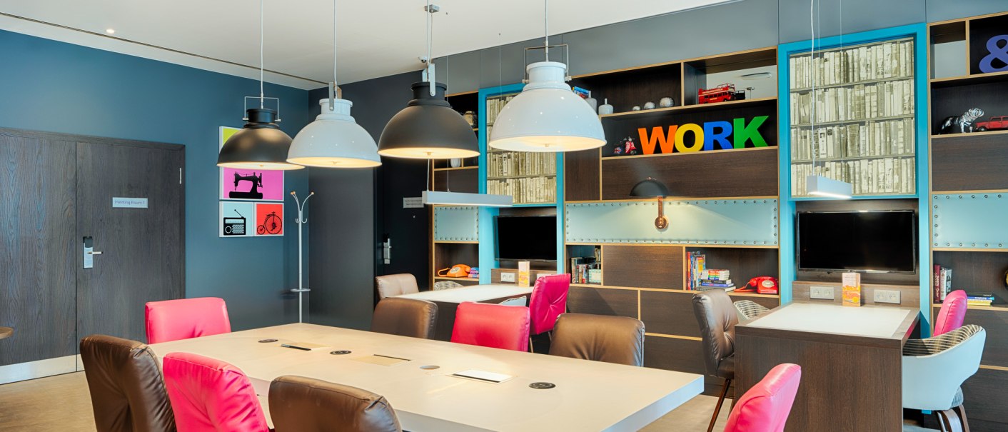 Moderner Arbeitsbereich mit bunten St&uuml;hlen, gro&szlig;en Tischen und Regalen. Helle Lampen h&auml;ngen von der Decke, im Hintergrund steht das Wort 'WORK' in bunten Buchstaben., &copy; Premier Inn Germany
