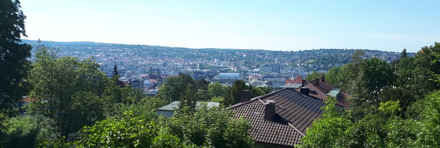Panoramic view of a city with green trees in the foreground and hills in the background under a clear sky., &copy; SMG