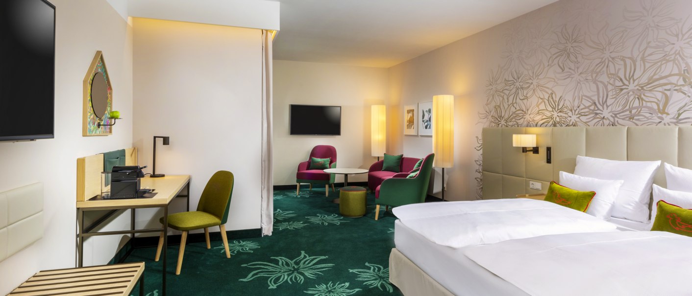 Moderne Hotelsuite mit Doppelbett, Schreibtisch, Sitzbereich und stilvoller Dekoration. Grüner Teppich und helle Wände mit floralen Mustern., © Mövenpick Hotel Stuttgart Messe & Congress