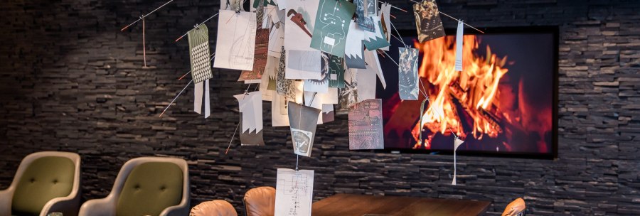 Moderner Raum mit Tisch, darüber hängen Papierblätter an Drähten. Im Hintergrund ein Bildschirm mit Kaminfeuer. Bequeme Stühle und Bücher auf dem Tisch., © TOMAS Moderner Raum mit Tisch, darüber hängen Papierblätter an Drähten. Im Hintergrund ein Bildschirm mit Kaminfeuer. Bequeme Stühle und Bücher auf dem Tisch., © TOMAS