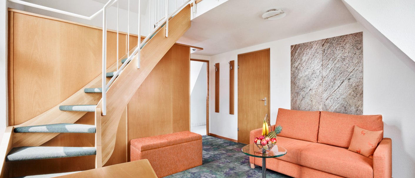 Moderne Junior Suite mit Treppe, orangefarbenem Sofa, kleinem Glastisch und frischem Obst. Helle Holzelemente und Teppichboden., © Hirsch Hotel