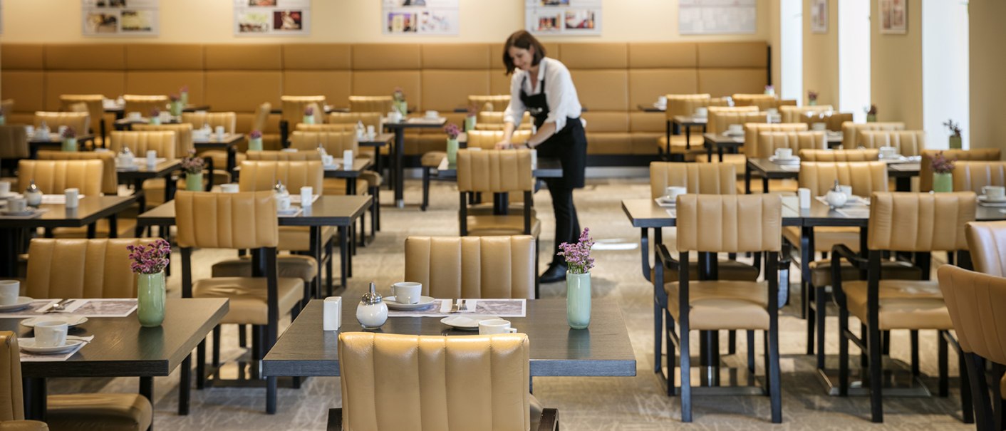 Ein Restaurant mit beige gepolsterten Stühlen und gedeckten Tischen. Eine Person richtet einen Tisch her., © Arcotel Hotels