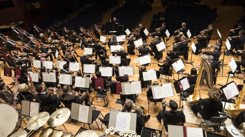 Ein Orchester spielt auf einer B&uuml;hne, Musiker mit verschiedenen Instrumenten, darunter Streicher und Bl&auml;ser, dirigiert von einer Person in der Mitte., &copy; Sebastian Klein