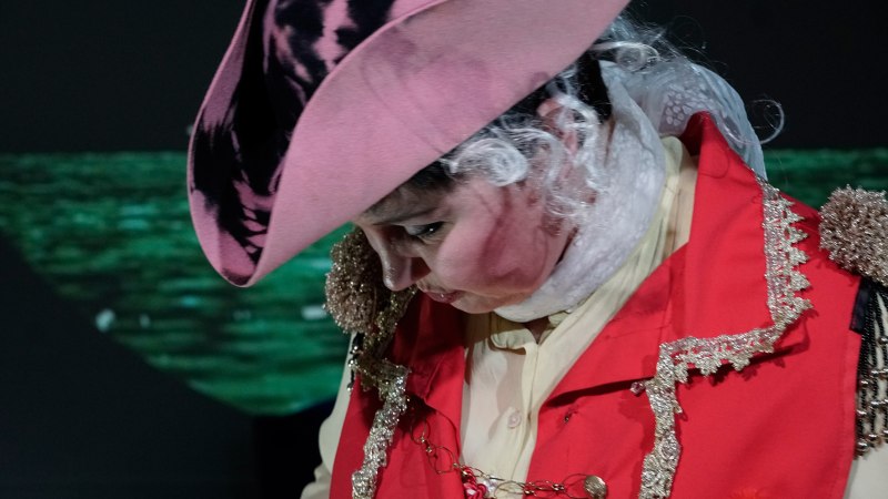 Eine Person in einem auff&auml;lligen historischen Kost&uuml;m mit rotem Mantel und rosa Hut, die nach unten schaut. Der Hintergrund ist dunkel und unscharf., &copy; Katja Ritter