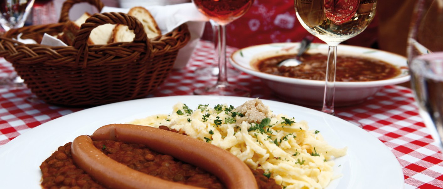 A plate of lentils, spaetzle and sausages on a red and white checkered tablecloth. A basket of bread and a glass of wine in the background., © Stuttgart-Marketing GmbH A plate of lentils, spaetzle and sausages on a red and white checkered tablecloth. A basket of bread and a glass of wine in the background., © Stuttgart-Marketing GmbH