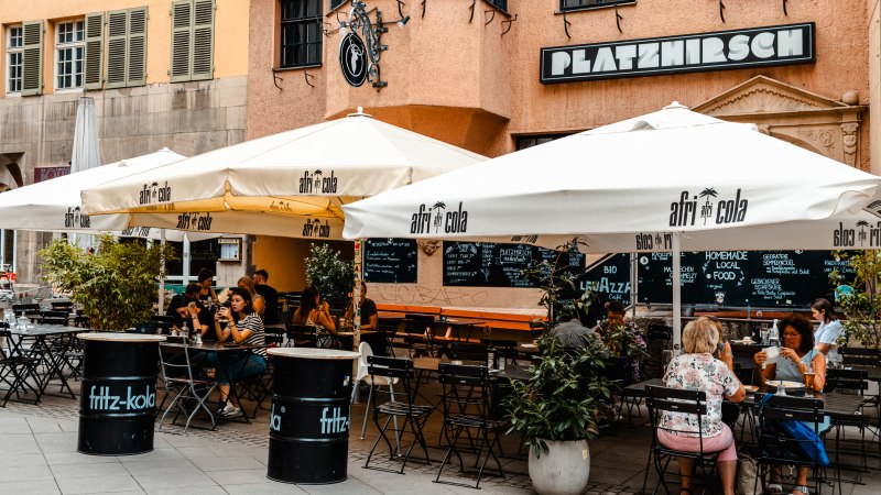 Außenbereich des Cafés Platzhirsch mit weißen Sonnenschirmen und Gästen an Tischen. Die Fassade ist orange, und es gibt Pflanzen als Dekoration., © SMG Stuttgart Marketing GmbH - Sarah Schmid