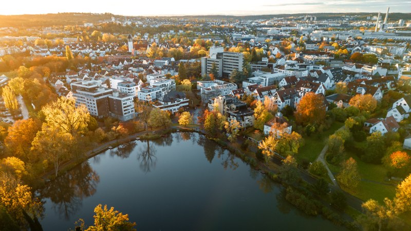 Aerial view of the Klostersee in Sindelfingen, surrounded by autumnal trees and residential buildings at sunset., © Christoph Partsch Aerial view of the Klostersee in Sindelfingen, surrounded by autumnal trees and residential buildings at sunset., © Christoph Partsch