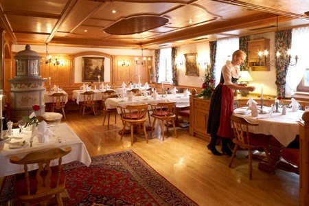 A cozy restaurant with wooden furniture, carpeting, and set tables. A server in traditional attire is serving at a table., © Jeanette Bender A cozy restaurant with wooden furniture, carpeting, and set tables. A server in traditional attire is serving at a table., © Jeanette Bender