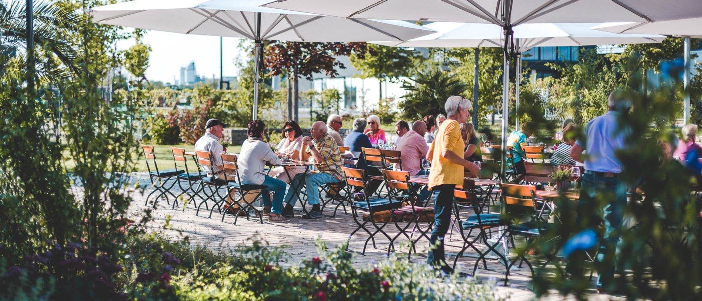 Menschen sitzen unter großen Sonnenschirmen an Holztischen im Freien, umgeben von grüner Vegetation und Blumen., © Wilhelm Kern GmbH Menschen sitzen unter großen Sonnenschirmen an Holztischen im Freien, umgeben von grüner Vegetation und Blumen., © Wilhelm Kern GmbH