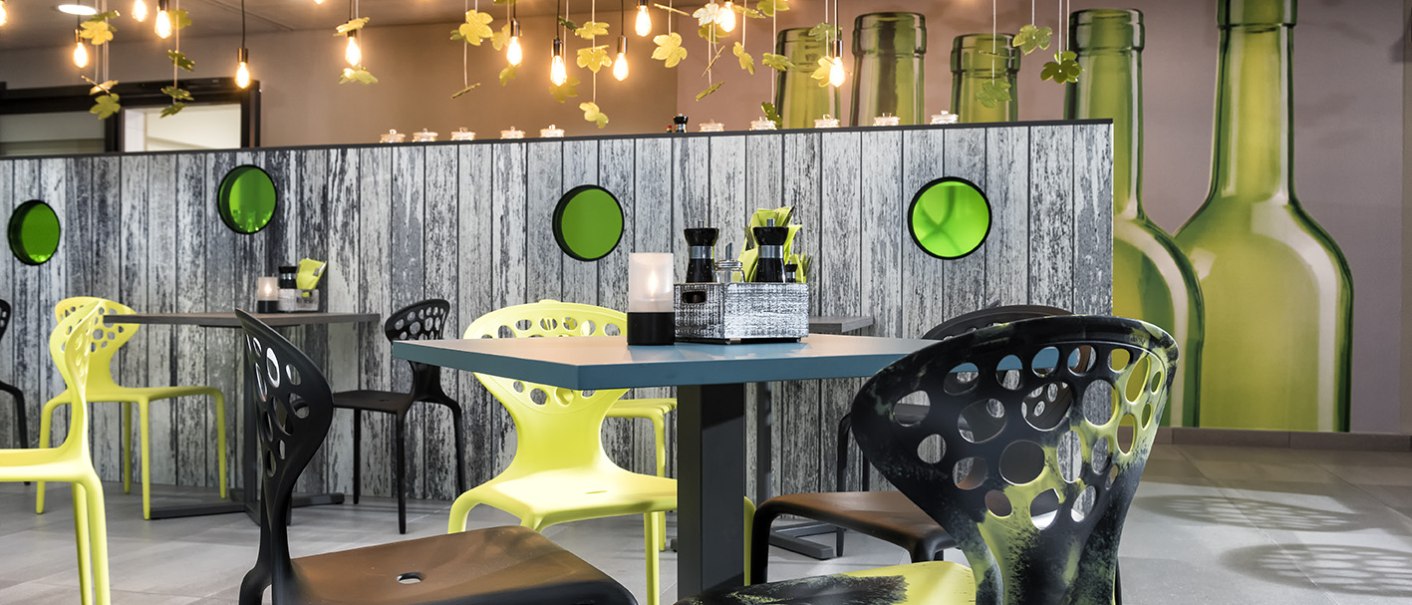 Modernes Restaurant mit gelben und schwarzen Stühlen, hängenden Flaschen und Blumen als Dekoration. Holzwand mit grünen Kreisen im Hintergrund., © ibis Styles Stuttgart Vaihingen Modernes Restaurant mit gelben und schwarzen Stühlen, hängenden Flaschen und Blumen als Dekoration. Holzwand mit grünen Kreisen im Hintergrund., © ibis Styles Stuttgart Vaihingen