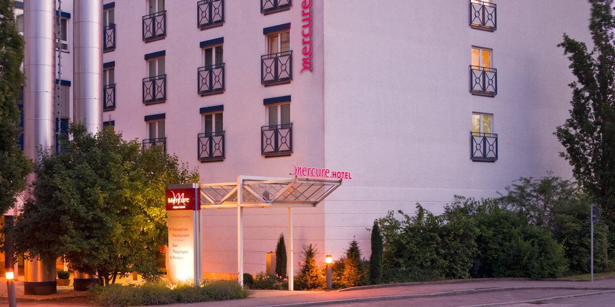 Exterior view of a Mercure Hotel with illuminated entrance, surrounded by trees and bushes., &copy; Mercure Stuttgart Airport Messe