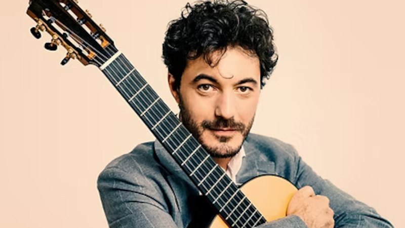 Ein Mann mit dunklem Haar h&auml;lt eine klassische Gitarre und schaut in die Kamera. Er tr&auml;gt einen grauen Anzug vor einem neutralen Hintergrund., &copy; Staatliche Hochschule f&uuml;r Musik und Darstellende Kunst