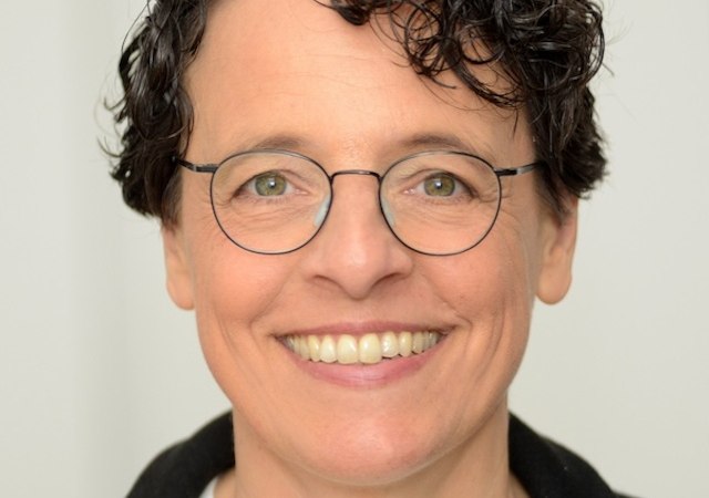 A portrait of a person wearing glasses and with curly hair, smiling warmly at the camera. The background is neutral and bright., © Renitenztheater Stuttgart e.V. A portrait of a person wearing glasses and with curly hair, smiling warmly at the camera. The background is neutral and bright., © Renitenztheater Stuttgart e.V.