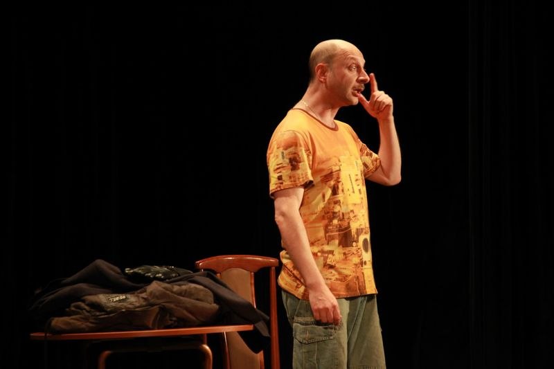 Ein Mann in einem orangefarbenen T-Shirt steht auf einer Bühne und gestikuliert. Neben ihm steht ein Tisch mit Kleidung., © Theater Atelier Ein Mann in einem orangefarbenen T-Shirt steht auf einer Bühne und gestikuliert. Neben ihm steht ein Tisch mit Kleidung., © Theater Atelier