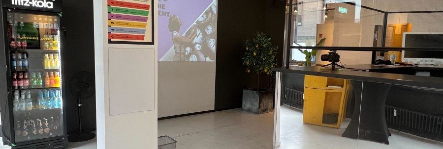 A room with a fridge, projector and reception desk. An introductory poster hangs on the wall. The room looks modern and inviting., &copy; pause & play Stuttgart