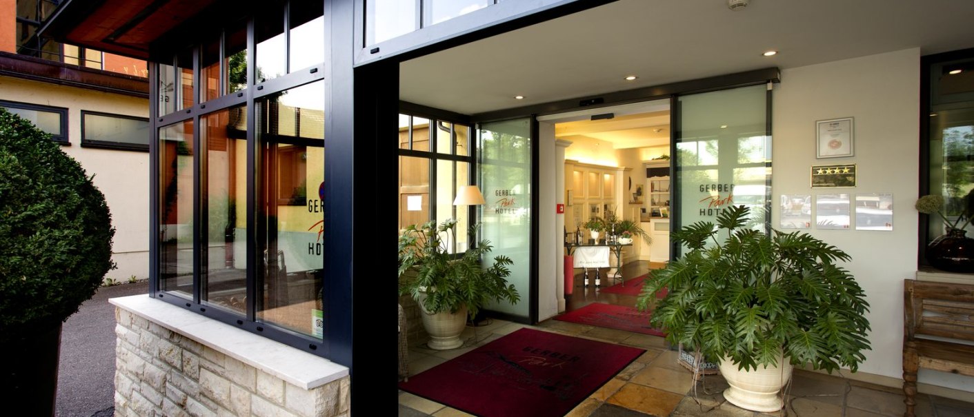 Moderner Hoteleingang mit Glasfront, roten Teppichen und großen Pflanzen. Innenbereich mit heller Beleuchtung sichtbar., © TOMAS Moderner Hoteleingang mit Glasfront, roten Teppichen und großen Pflanzen. Innenbereich mit heller Beleuchtung sichtbar., © TOMAS