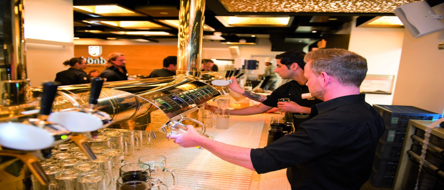 Barkeeper zapfen Bier an einer glänzenden Theke. Im Hintergrund sind weitere Personen und eine stilvolle Decke zu sehen., © Schönbuchbrauerei, Sebastian Berger