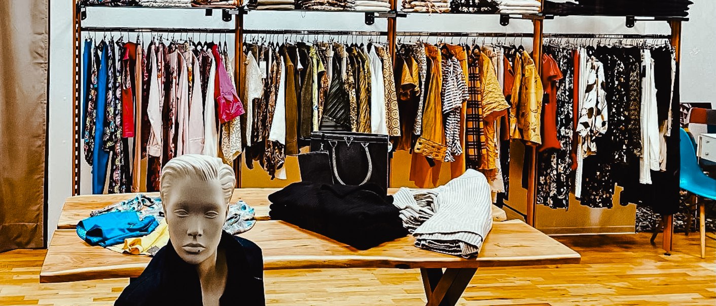 Ein Bekleidungsgeschäft mit Kleiderständern, gefalteten Kleidungsstücken und einer Schaufensterpuppe. An der Wand hängen Modefotos., © StuttgARTfactory