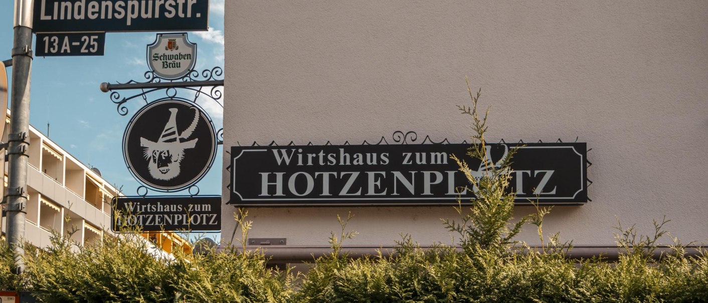 Schild des Wirtshauses zum Hotzenplotz an der Lindenspürstraße, umgeben von grünen Büschen und einem blauen Himmel im Hintergrund., © SMG. Sarah Schmid