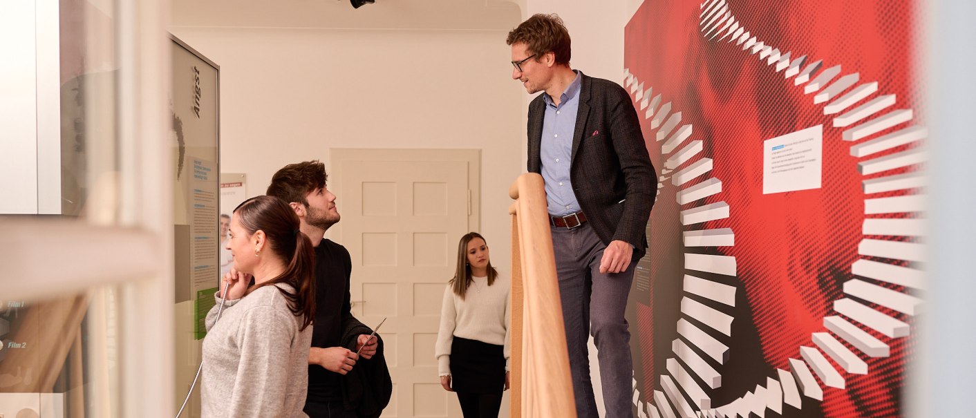 Menschen in einem Ausstellungsraum. Eine Person steht auf einer Treppe, andere betrachten eine Wand mit rotem Design. Thema: Angst., © Stiftung Psyche